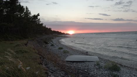 Baltic-seashore-during-sunset,-waves-hitting-the-rocky-beach-with-a-small-rowboat-on-the-rocks,-wind-moving-plants-and-trees