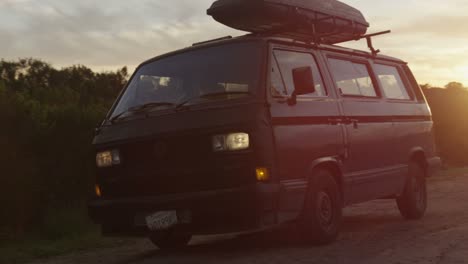 a surfer pulls up in a camper van with a board on top at daybreak on a dirt road in a coastal area