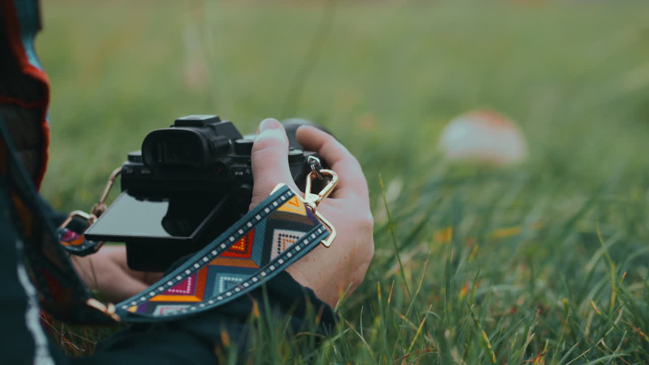 Close up of a Camera with colourful strap and woman's hands photographing mushroom in the grass in nature during a cold windy day in slow motion