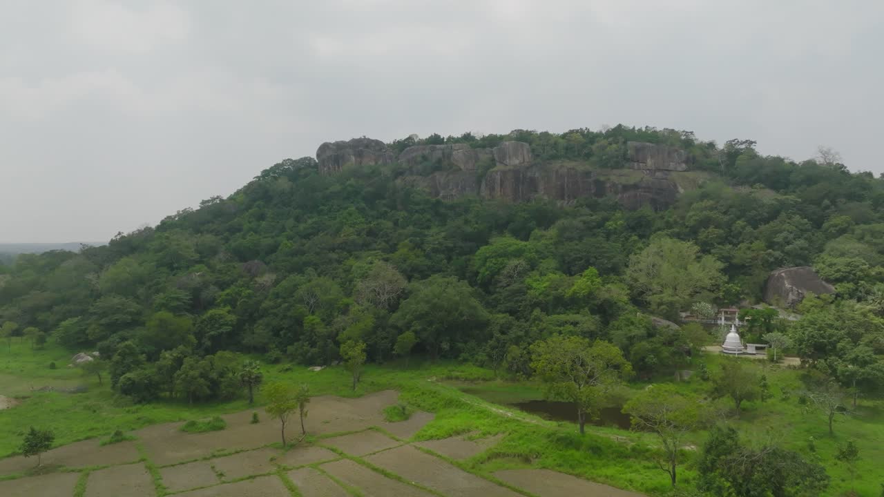 Drone footage showcasing a green forest hill with rocky cliffs and farmland below under cloudy skies