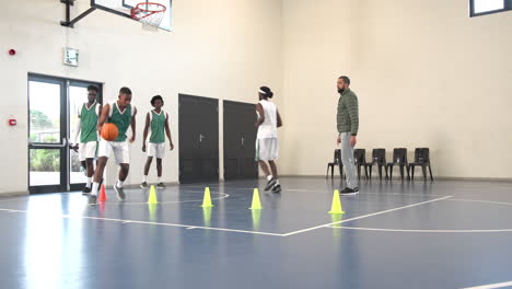 multiracial male basketball players bouncing basketball and avoiding cones with coach in gym