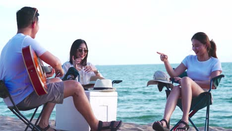 happy young people enjoying with music at the beach, young man playing guitar and singing as his friends clapping and laughing, having fun on the beach picnic in summer holidays, vacation.