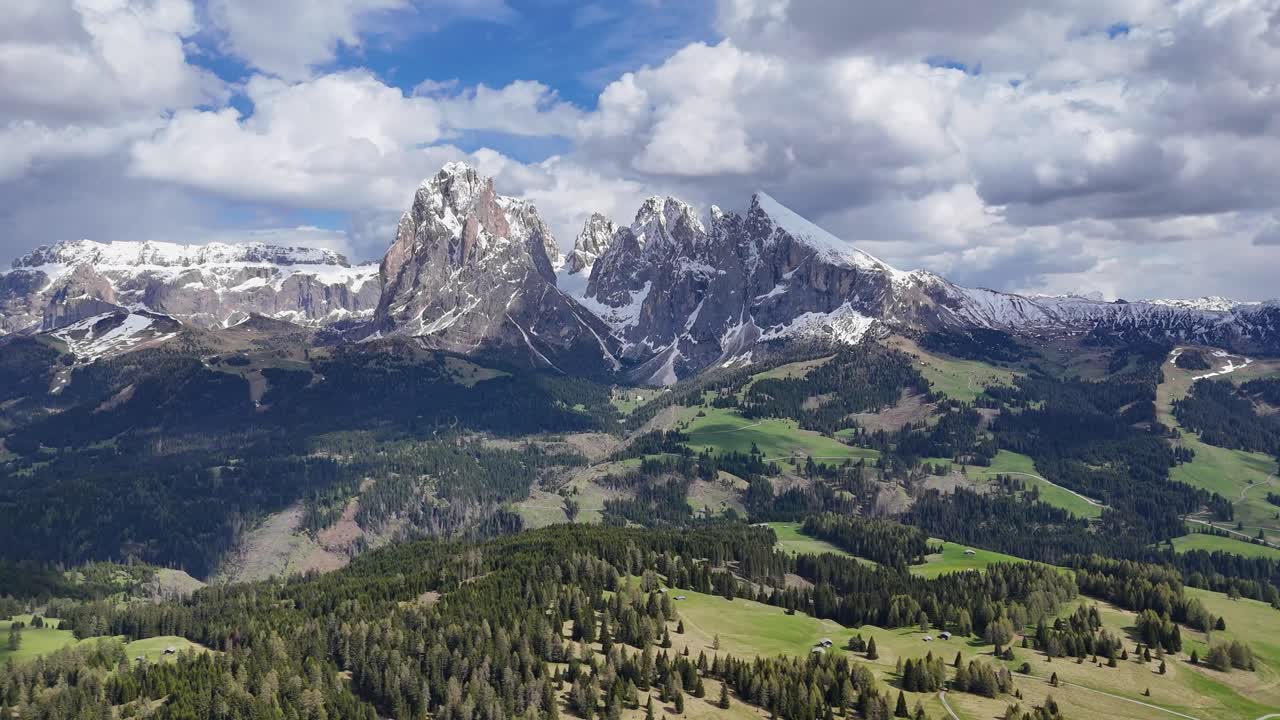Drone footage of Alpe di Siusi shows Sassolungo Group, ascending forward over pine forests and rolling hills under a partly cloudy sky