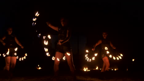 Tres-Mujeres-Con-Aros-Encendidos-Bailan-Con-Antorchas-Ardientes-Vestidas-De-Cuero-En-Un-Hangar-Oscuro-Demostrando-Un-Espectáculo-De-Fuego-De-Circo-En-Cámara-Lenta