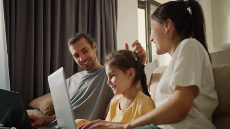 Family-sitting-on-the-sofa-and-looking-at-something-on-a-gray-laptop.-A-brunette-girl-in-a-white-T-shirt,-her-daughter,-a-girl-in-a-yellow-dress,-and-a-brunette-man-in-a-gray-T-shirt-are-sitting-together-on-a-light-brown-sofa-and-looking-at-the-screen-of-a-gray-laptop-in-a-modern-apartment