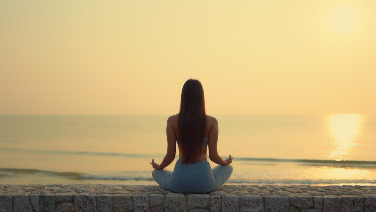 Toma trasera y amplia de una mujer asiática delgada meditando durante un amanecer dorado sobre el océano