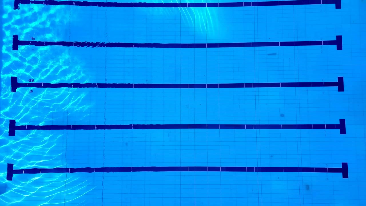 Vista aérea calma de uma piscina interior vazia com raias paralelas, água azul, marcações escuras, piso de azulejos e reflexos de luz cintilantes criando.