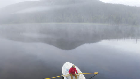 aerial view of going over a person rowing a boat on a foggy lake with forest and mountain in the landscape