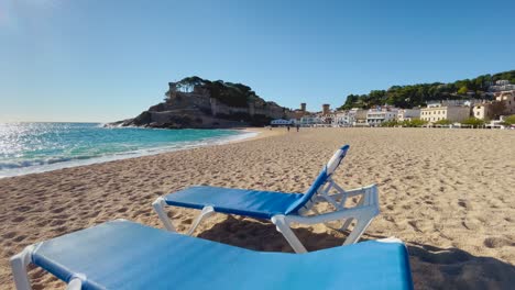 Deck-chairs-on-the-beach-with-the-background-of-walled-castle-over-the-sea-in-Tossa-de-Mar,-Girona-Spain-Costa-Brava-turquoise-water-beaches