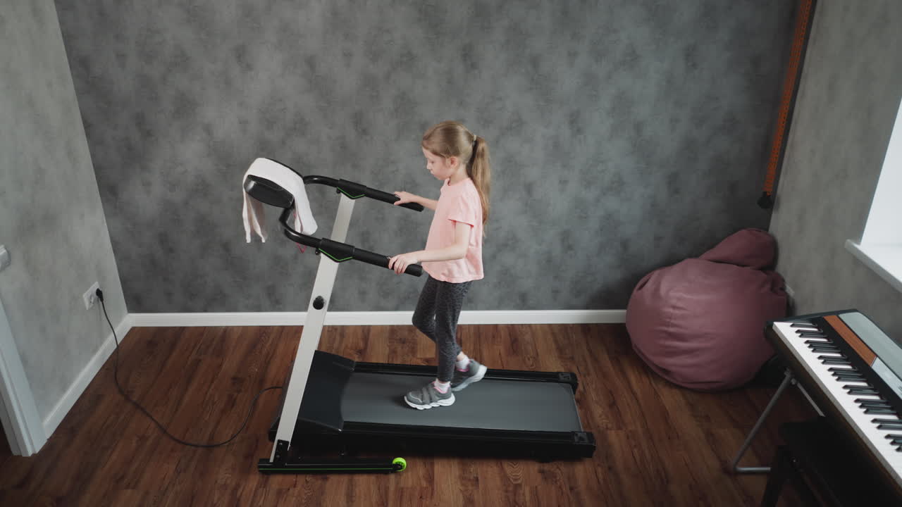 Top down view of teenage girl in pink shirt and patterned leggings walking on treadmill holding handles in home workout room with textured grey wall wooden floor partial piano and beanbag visible