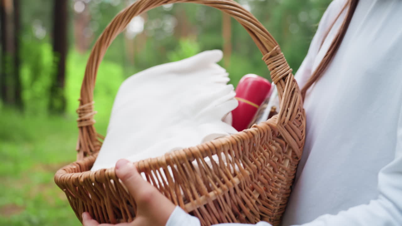Primer plano de una chica que lleva una cesta de pícnic con ambas manos mientras camina por un sendero verde en un bosque en un tranquilo día de verano, mostrando detalles de la manta y la botella, que simboliza el ocio y un momento de aventura al aire libre.