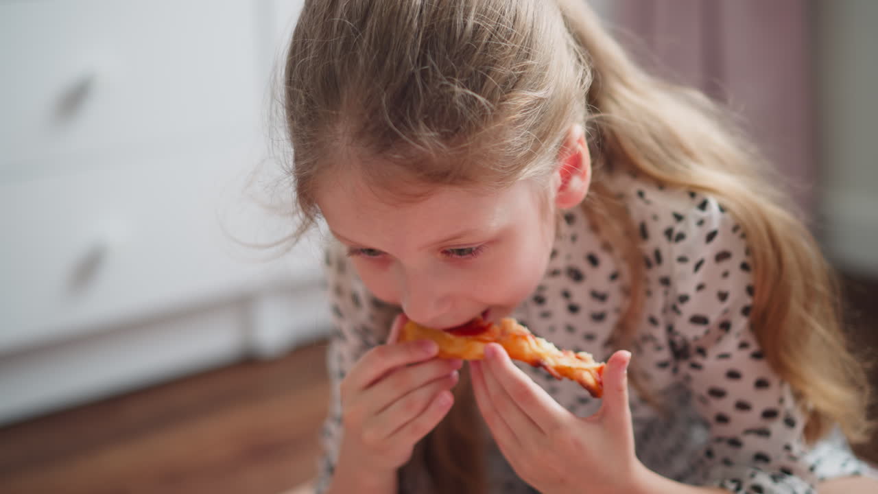 Young girl in spotted outfit bent over eating slice of cheesy pepperoni pizza indoors, close up view, focused on food, wardrobe blurred in background