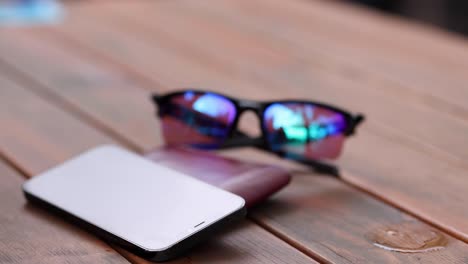 a close-up of sunglasses and a smartphone on a wooden table with a hand holding a beer glass.