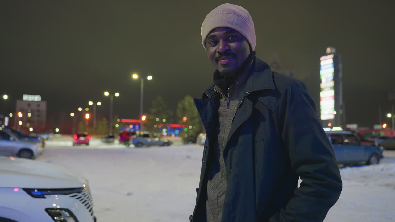 Un autista con cappello e cappotto invernali posa con sicurezza in un parcheggio innevato di notte, con auto e luci della città sullo sfondo; ritratto illuminato da lampioni e bagliori colorati