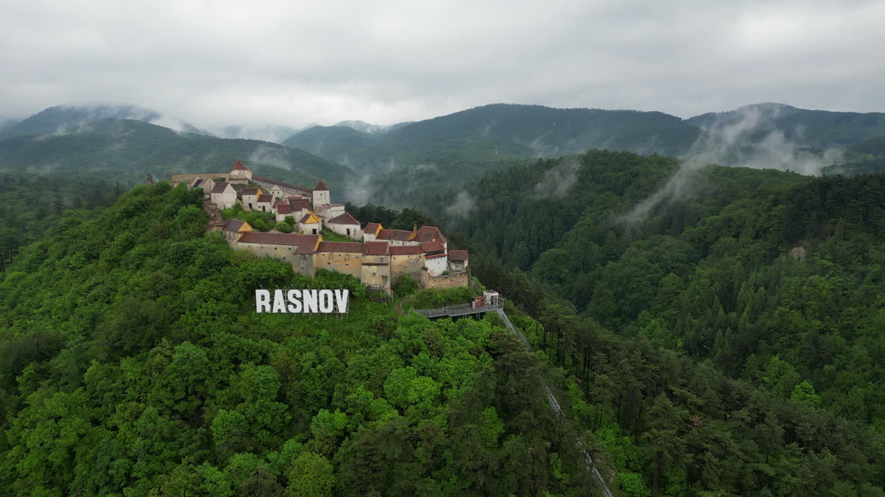 Majestic Rasnov Citadel perched atop a hill, surrounded by lush green forest, in a cloudy day