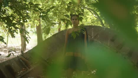 A-young-woman-of-Indian-beauty,-dressed-in-an-elegant-eco-friendly-dress,-stands-on-an-old-boat-amidst-trees-and-green-leaves