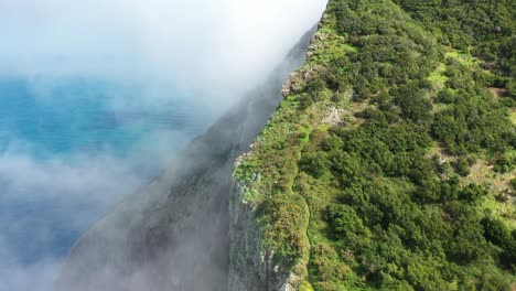 thin clouds are moving slowly over the cliff on the coast of madeira island with the turquoise and deep blue water below and green lush nature on the top with a small trail leading up the mountain