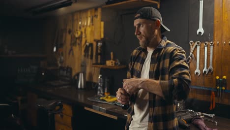 happy guy mechanic drinks soda from a glass bottle after finishing his working day in a cozy workshop studio