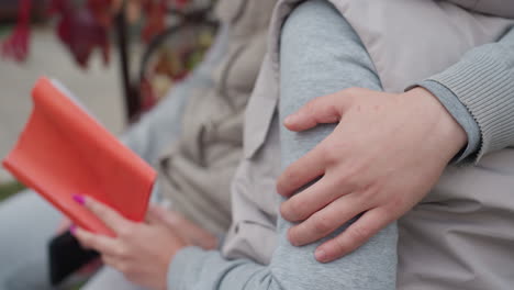 close up of hand warmly rubbing person arm while seated outdoors, as another person holds open book with bright orange cover, capturing moment of affection