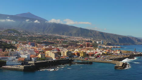 Malerische-Aussicht-Auf-Die-Pulsierende-Stadt-Und-Den-Hafen-Von-Puerto-De-La-Cruz,-Kanarische-Inseln,-Spanien,-An-Der-Küste-Des-Atlantischen-Ozeans,-Pico-De-Teide-Im-Hintergrund,-Blaues-Meer-Und-Himmel,-Luftaufnahme-4k