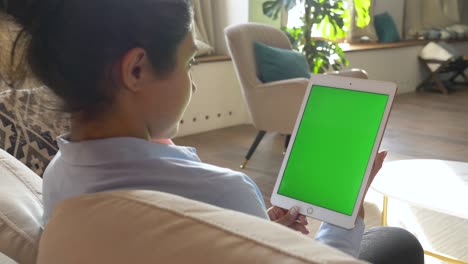 vista por encima del hombro de la mujer india utiliza una tableta con pantalla verde y seguimiento de movimiento. niña sentada en el sofá en casa y tocando o desplazando la página de noticias en el gadget con la tecla chroma de cerca