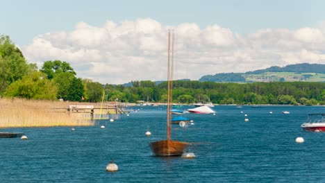 a classic wooden sailboat on lake murten, fribourg, switzerland, surrounded by calm waters and lush green landscapes, captured in a relaxing timelapse. the sailboat sways in the wind at anchor.