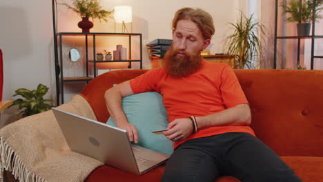 young bearded man using credit bank card and laptop, transferring money, purchases online shopping