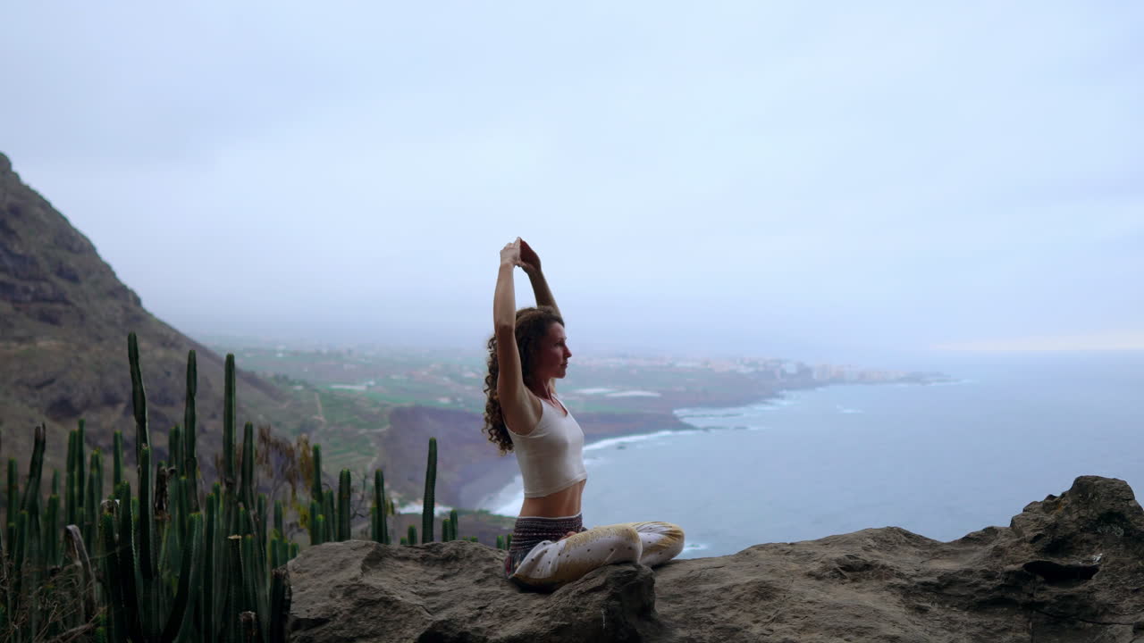 Atop a mountain, a woman meditates and showcases the Maha Sakal hand gesture against the ocean and the expanse of green mountains