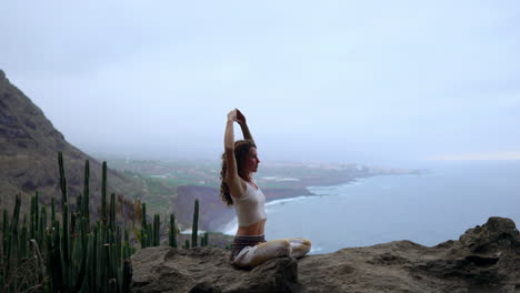en la cima de una montaña, una mujer medita y muestra el gesto de la mano maha sakal contra el océano y la extensión de las montañas verdes
