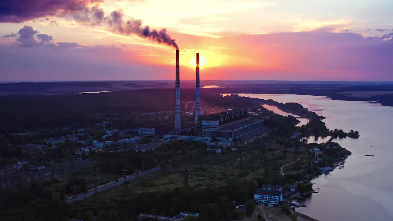 Industrial zone in the evening nature. Dark smoke pours out of factory chimney at sunset. Factory near the river against background of thick clouds. Environmental pollution.