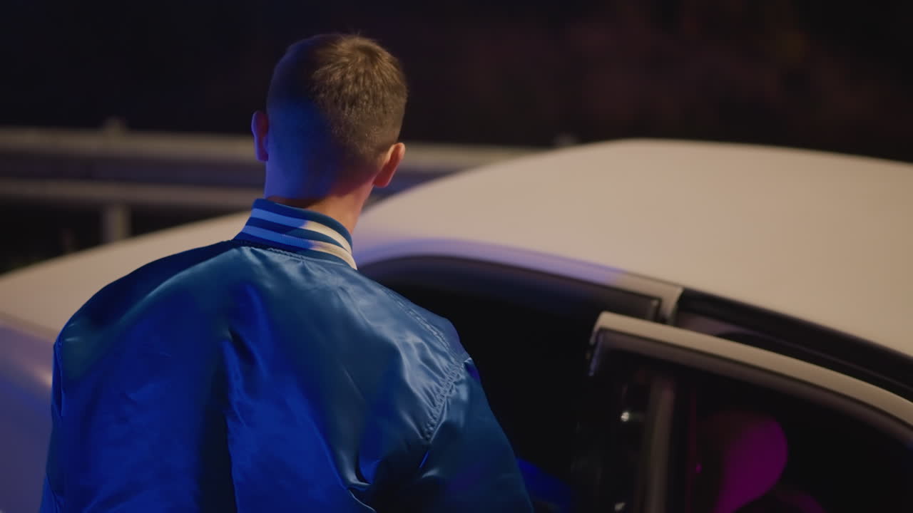 Giovane guidatore che cammina verso l'auto in un parcheggio vuoto, giubbotto blu da studente che cattura la luce del lampione, ispeziona la portiera e scruta l'interno illuminato al neon, esita prima di entrare; l'inquadratura cinematografica aumenta l'attesa.
