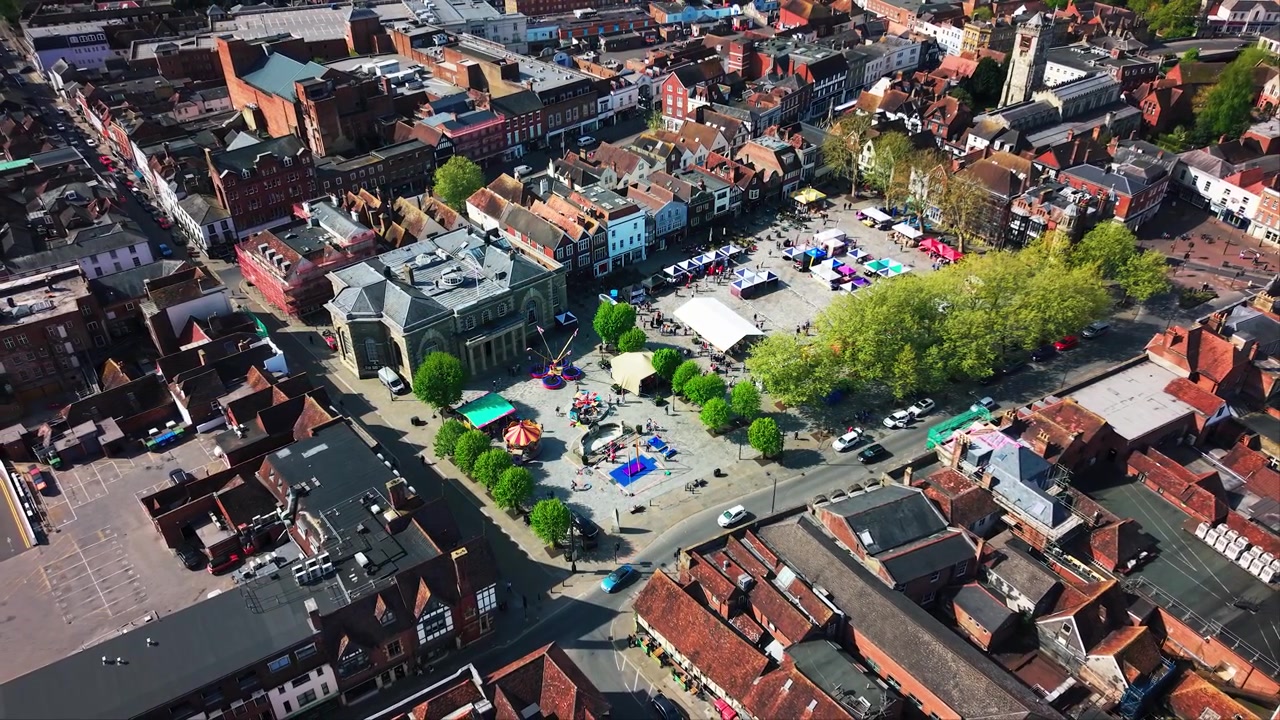 Drohnenaufnahme, die sich über den Marktplatz von Salisbury während einer Veranstaltung zum St. Georgstag zurückzieht und die Guildhall, Marktstände, Fahrgeschäfte, Menschenmengen, Dächer und Straßen zeigt