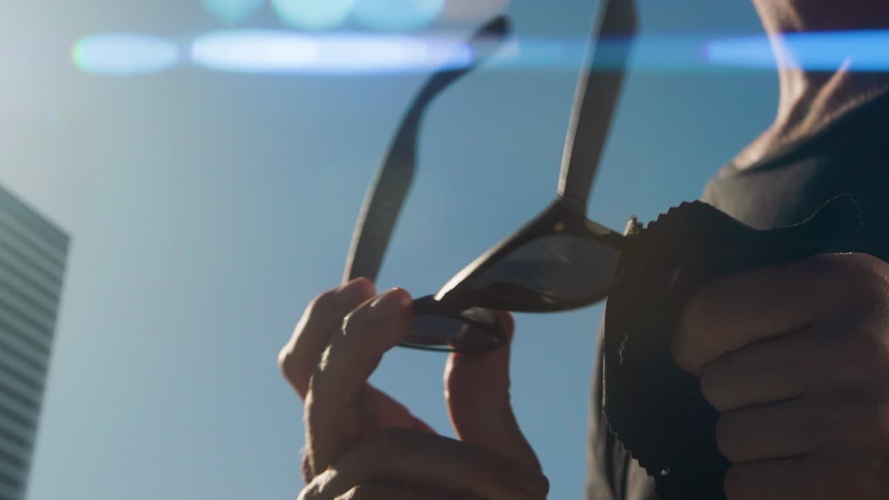 검은 선글라스의 왼쪽 렌즈를 는 손, 검은 티셔츠를 입은 남자의 몸통, 푸른 하늘과 배경의 건물, 빛의 아나모르픽 발광