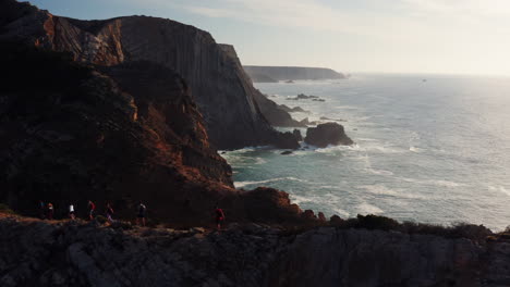 aerial: silhouette of hiker group walking on narrow trail with beautiful coastline with crashing atlantic ocean during sunset