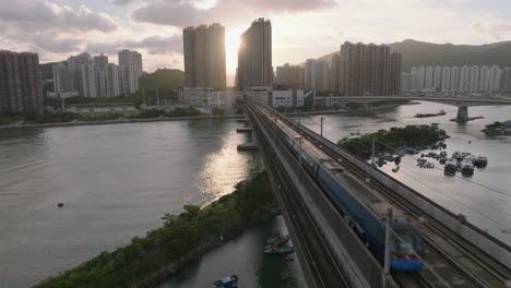 Aerial-backwarrds-shot-revealing-airport-express-train-on-bridge-towards-Tsing-Yi-during-golden-hour-sunset,-sun-flares-in-the-background