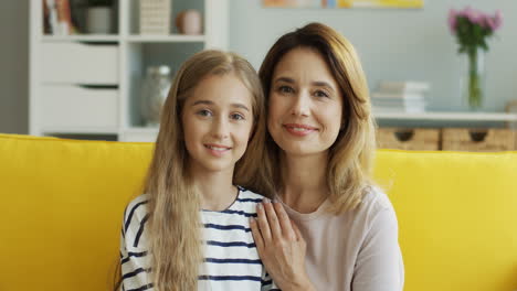 Retrato-De-Madre-Rubia-E-Hija-Adolescente-Sentadas-En-Un-Sofá-Amarillo-Y-Mirando-A-La-Cámara-Mientras-Sonríen