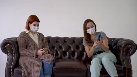 two women wearing medical face mask, one shows the other something funny on the phone. females sitting on a sofa.