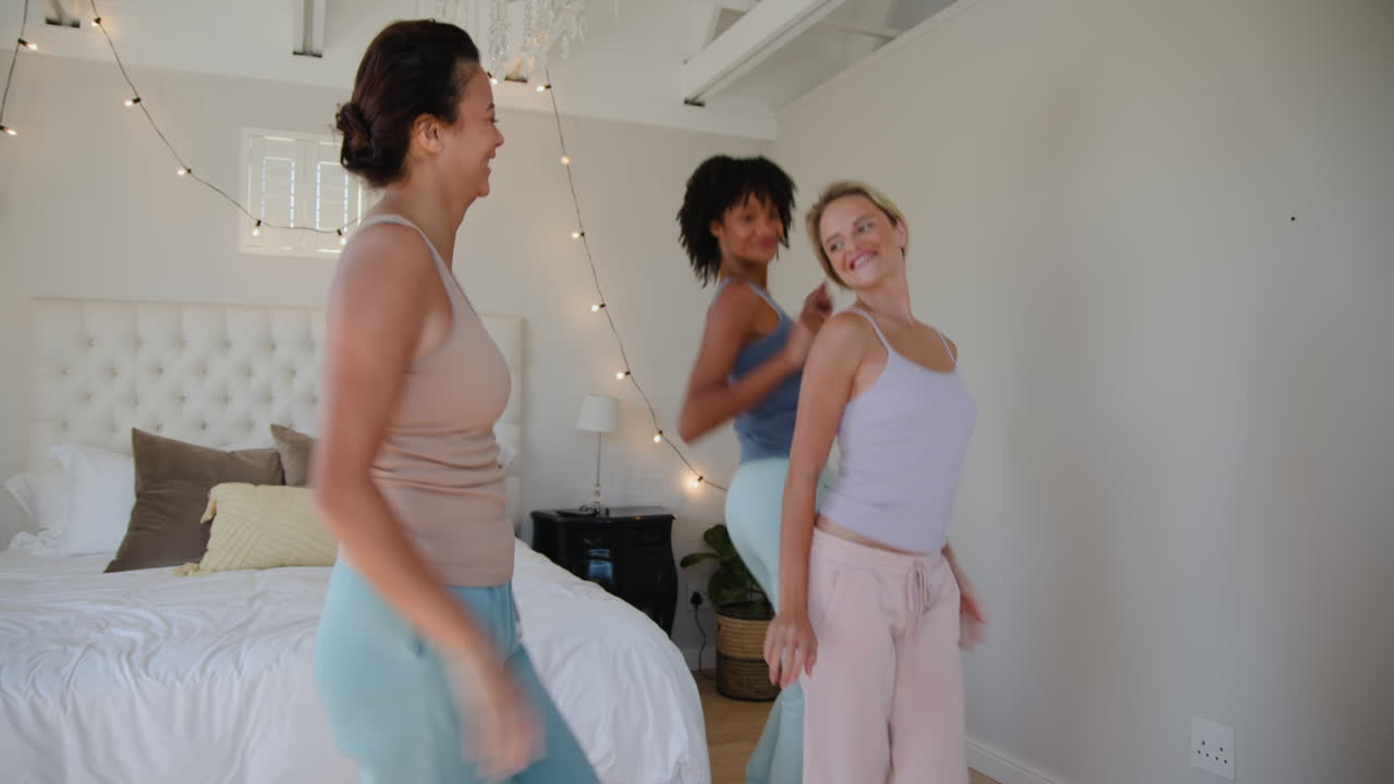 Diverse female friends dancing joyfully in bedroom, enjoying fun and laughter together, at home