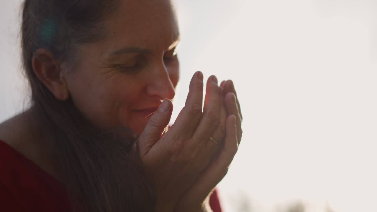 Donna che tiene in mano foglie di menta, sfondo di campo soleggiato con calda retroilluminazione mentre inala il profumo, momento intimo di consapevolezza e calma, sorriso gentile e postura rilassata che suggeriscono un rituale di aromaterapia.