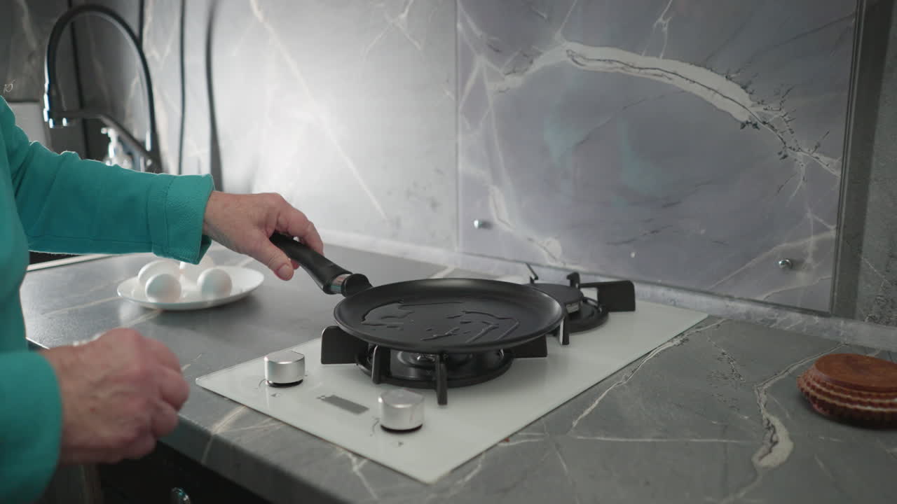 Elderly woman in green sweatshirt cooking on stove, flipping pancake in pan. Fresh eggs on countertop, modern kitchen setting with sleek design, showcasing independence and healthy cooking