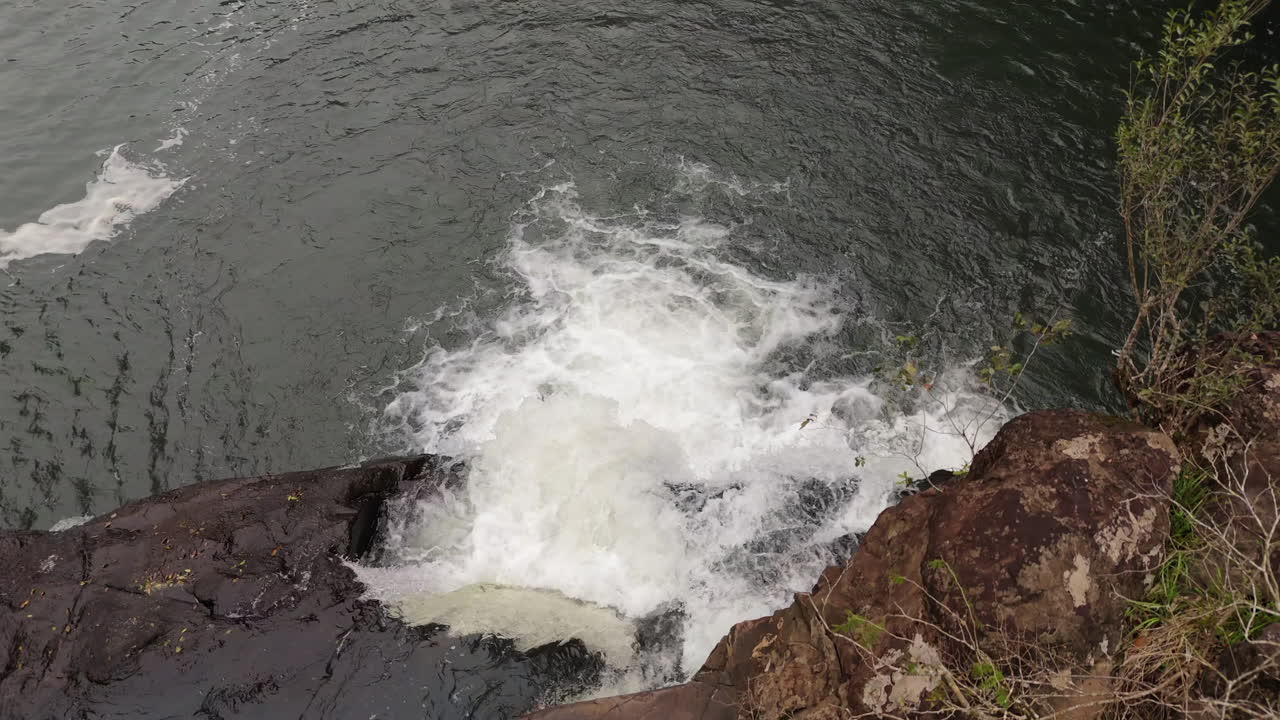 Close-up drone view of water rushing over rocky edge creating white foam waterfall splash.