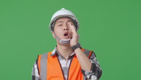 close up of asian male engineer with safety helmet yelling with hand over mouth while standing in the green screen background studio