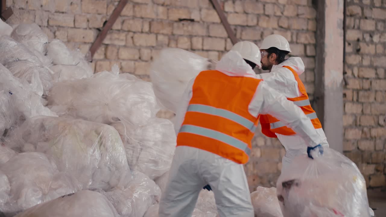 un hombre de piel negra en un uniforme de protección blanco un chaleco naranja y un casco blanco apila bolsas de celofán y polietileno en una gran pila junto con su colega en una gran planta de procesamiento y clasificación de residuos