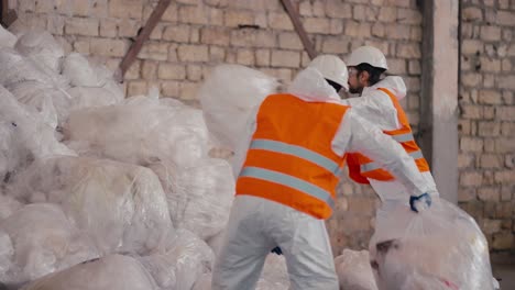 Un-Hombre-De-Piel-Negra-Con-Un-Uniforme-De-Protección-Blanco,-Un-Chaleco-Naranja-Y-Un-Casco-Blanco-Apila-Bolsas-De-Celofán-Y-Polietileno-En-Una-Gran-Pila-Junto-Con-Su-Colega-En-Una-Gran-Planta-De-Procesamiento-Y-Clasificación-De-Residuos.