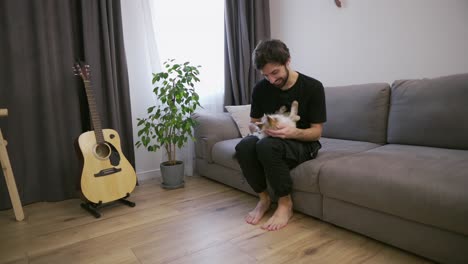 Un-Joven-Sentado-En-Un-Sofá-Acaricia-A-Un-Gato-Atigrado-De-Rodillas.