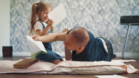 playful girl with ponytails hits dad with notebook on floor