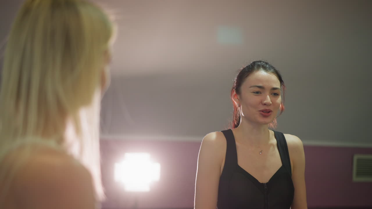 scena in palestra con un'allieva asiatica che parla con l'allenatrice, due donne in studio illuminate da un caldo controluce, l'atleta con reggiseno sportivo nero riceve istruzioni dalla trainer bionda, dialogo di supporto su tecnica e forma