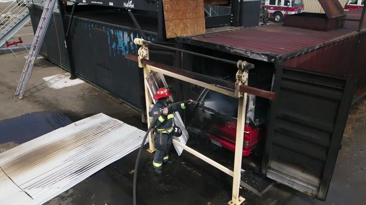 imágenes aéreas de drones en cámara lenta de un bombero con una chaqueta de casco de equipo completo extinguiendo el fuego en un camión en un contenedor de metal que se desplaza sobre los servicios de emergencia de agua y mangueras