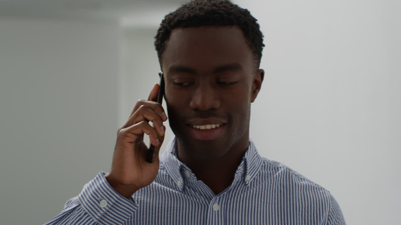 Close Up Of Smiling Young Businessman Walking Along Corridor In Office Talking On Mobile Phone