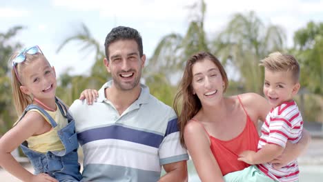 Portrait-of-happy-caucasian-parents-carrying-children-at-swimming-pool-at-beach-house